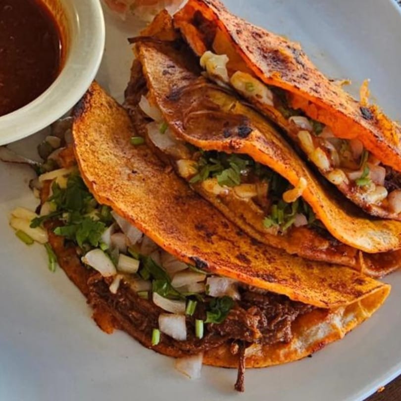 Three Agaveros birria tacos with chopped onions, cilantro, and a side of consommé.
