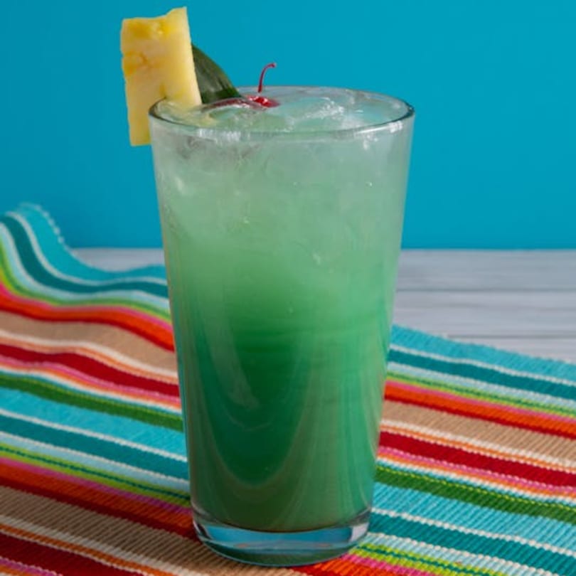 A vibrant green cocktail with pineapple and cherry garnish on a colorful striped placemat.