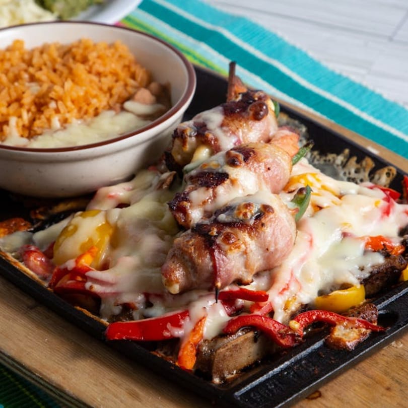Bacon-wrapped jalapenos with melted cheese, bell peppers, and a side of Mexican rice on a hot skillet.