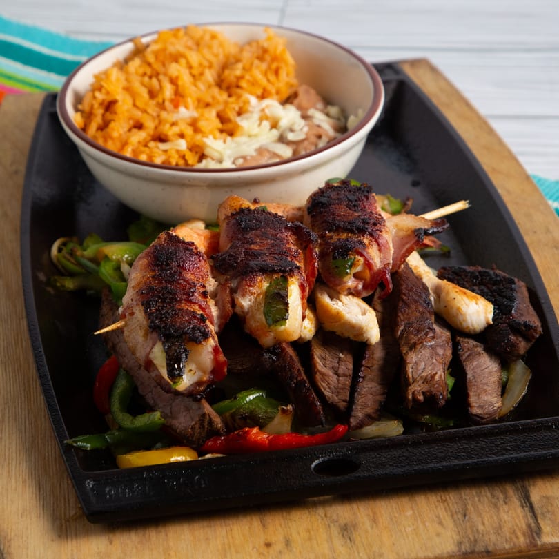 Sizzling fajitas with steak, bacon-wrapped shrimp, peppers, onions, and a side of rice and beans.
