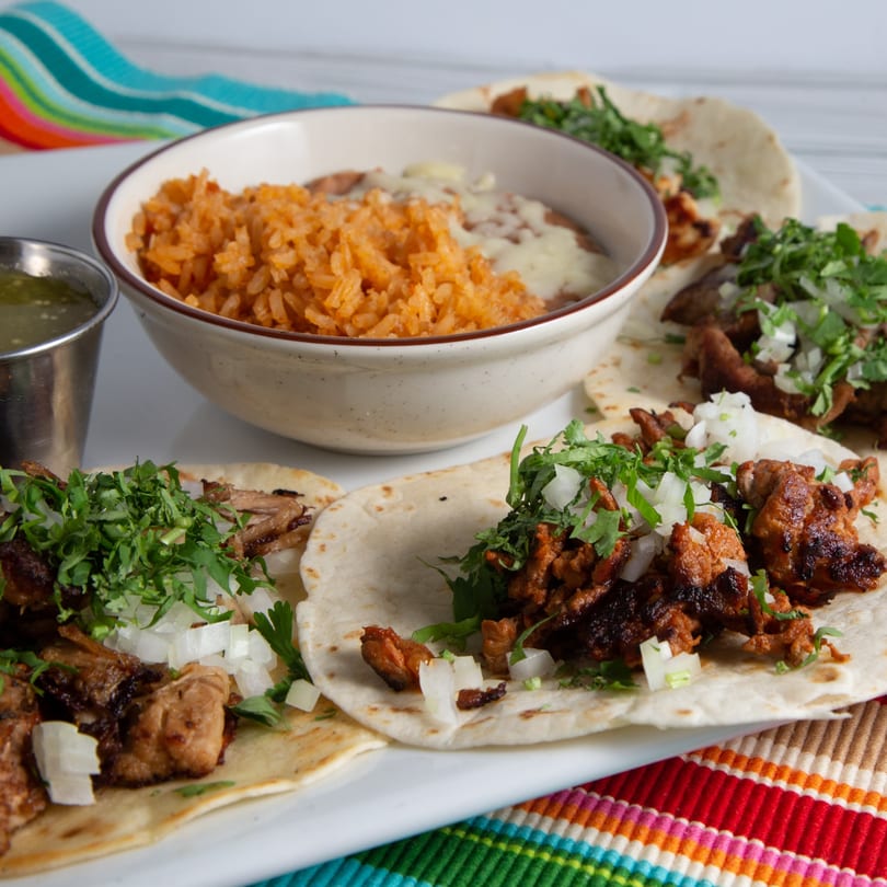 A plate of Agaveros Mexican Bar and Grill tacos with rice, beans, and salsa verde.