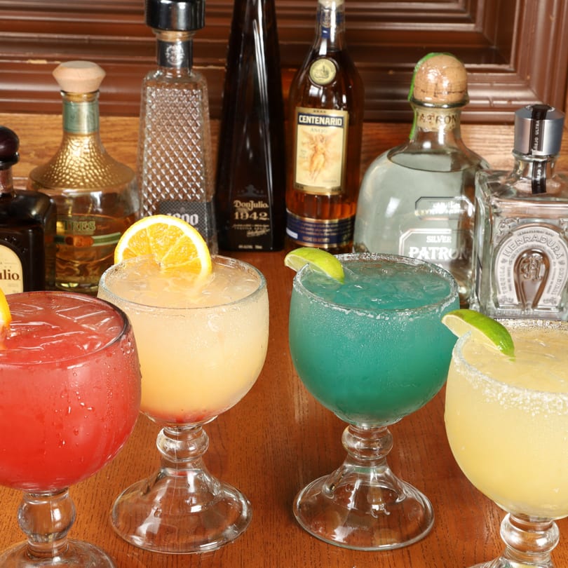 Four colorful margaritas in goblet glasses with tequila bottles in the background at Agaveros Mexican Bar and Grill.