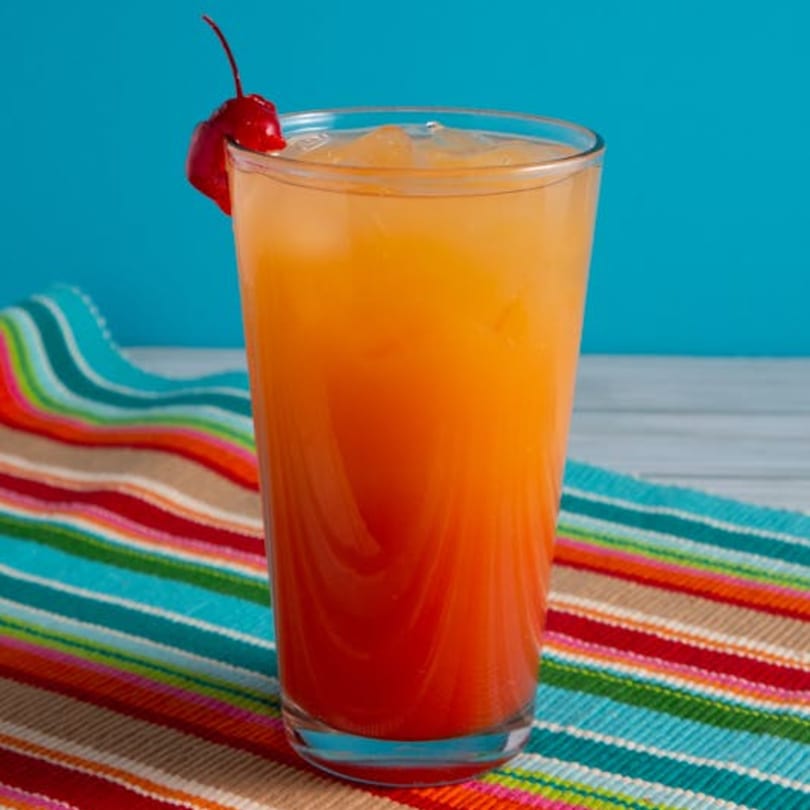 A vibrant orange and red Mexican sunrise cocktail with a cherry garnish on a colorful striped placemat.