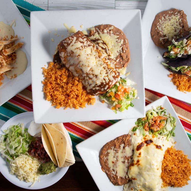 A variety of Mexican dishes, including enchiladas, tacos, rice, beans, and fresh toppings, on a colorful striped placemat.