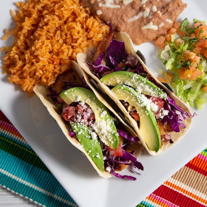Two shrimp tacos with avocado, purple cabbage, rice, refried beans, and a side salad.